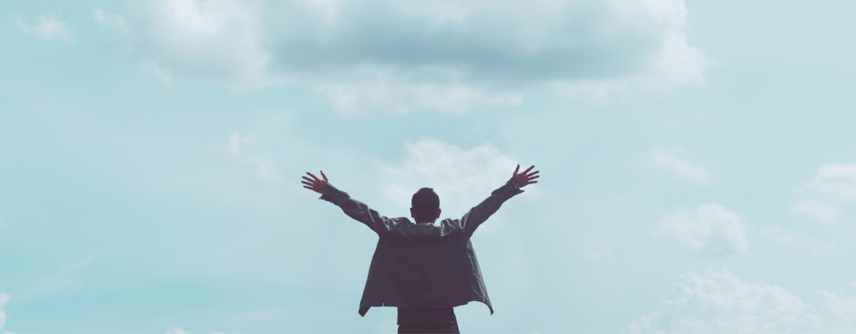 Une personne se tient debout, les bras tendus, face à un ciel nuageux, vu de dos. Le dégradé bleu-vert évoque le calme, comme si elle embrassait la liberté après une voiture tonneau, créant une atmosphère exaltante.